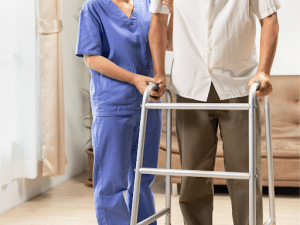 A nurse in blue scrubs assists an elderly man move with a walker