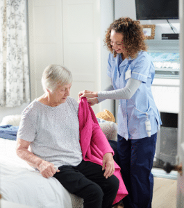 A female nurse helps an elderly woman put on a pink sweater at a nursing home