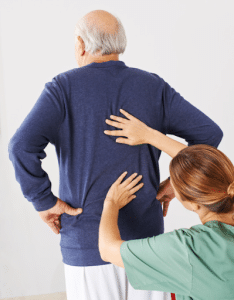 A female physical therapist wearing green scrubs examines an old man’s back