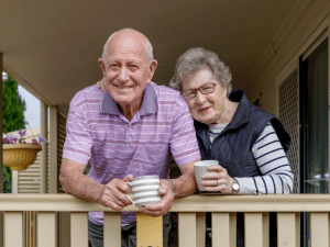 An elderly couple standing on their porch with coffee mugs in hand.
