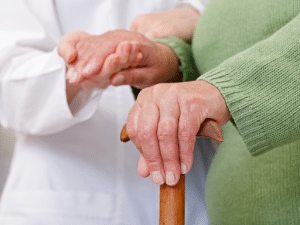 A doctor in a white lab coat holds the hand of an elderly woman in a green sweater with a cane
